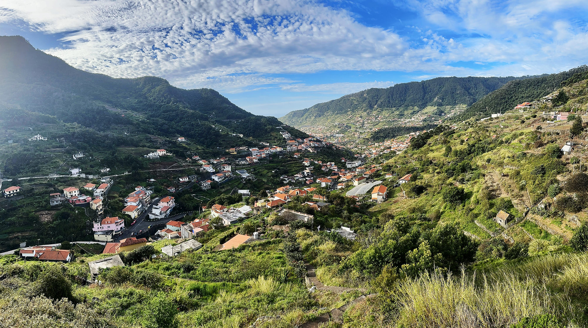 Panoramic view of a verdant valley on Madeira with scattered houses and terraced hillsides, leading towards the coast under a partly cloudy sky.