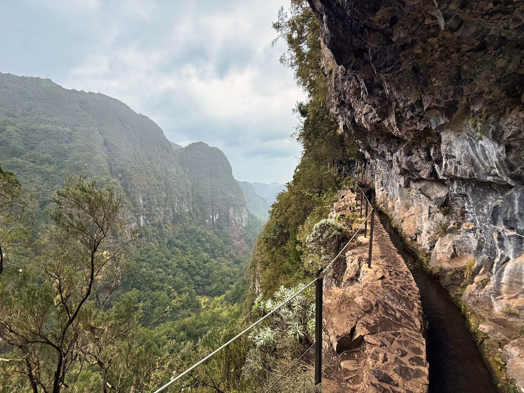 Úzká stezka Levada do Caldeirão Verde podél skalní stěny s výhledem do zeleného údolí.